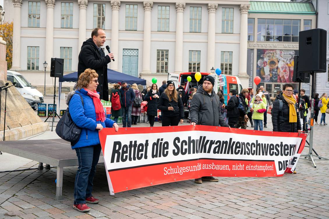 Ronny Kretschmer steht mit einem Mikrofon in den Händen auf einem Podest, davor stehen mehrere Personen mit einem Transparent mit der Aufschrift "Rettet die Schulkrankenschwestern! DIE LINKE."