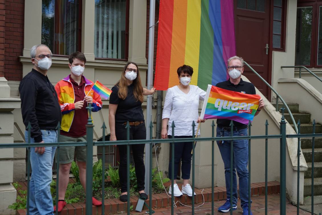 Stefan Wollenberg, Anton, Anja Mayer, Katharina Slanina und Carsten Schneider stehen vor einem fahnenmast, an dem eine Fahne in Regenbogenfarben hängt