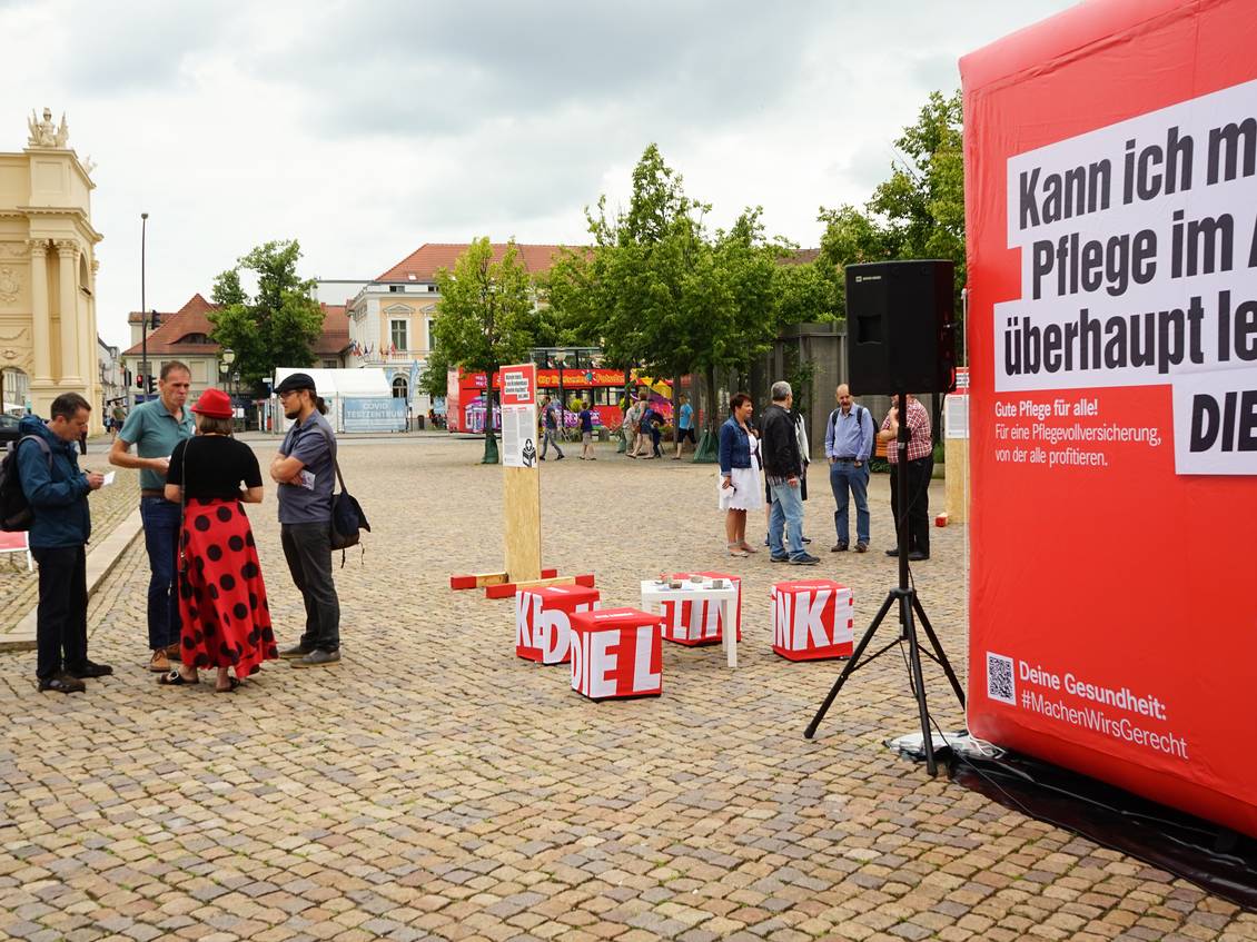 Luisenplatz in Potsdam, auf dem Platz steht ein 3x3 Meter großer aufblasbarer roter Würfel mit dem Logo der LINKEN und verschiedenen Aussagen zu den Themen Pflege und Gesundheit, um den Würfel herum stehen mehrere Menschen in Gruppen