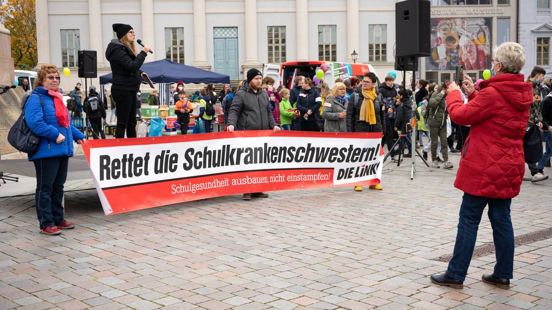 Anja Mayer steht mit einem Mikrofon in den Händen auf einem Podest, davor stehen mehrere Personen mit einem Transparent mit der Aufschrift "Rettet die Schulkrankenschwestern! DIE LINKE."
